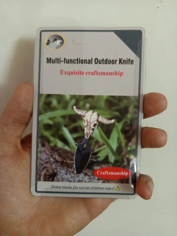 تصویر بستهبندی مقوایی-پلاستیکی یک چاقوی چندمنظوره فضای باز (Multi-functional Outdoor Knife) در دست یک نفر. روی کارت نوشتههای انگلیسی و یک تصویر از چاقوی مشکی با دسته جمجمه گاو دیده میشود. در پایین کارت، هشدار تیزی تیغه و دوری از کودکان به همراه یک مثلث